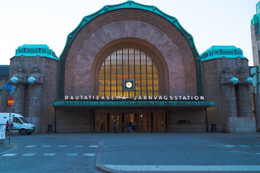 The façade of the central train station in Helsinki. A large arch in the middle, with two human statues holding globes on either side.