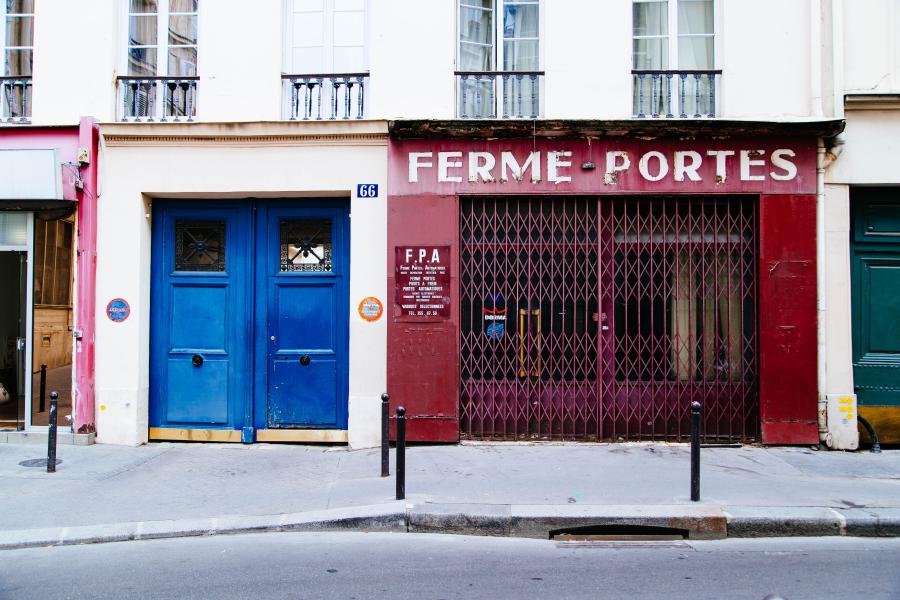 A red store front that reads "FERME-PORTES" has a closed gate. It is painted red, though shows many signs of age (deteriorating wood, broken signs, cracks). To its right is a clean set of blue doors to a residential building.